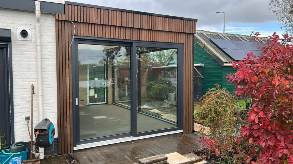 Uitbouw plaatsen met houten gevelbekleding en grote glazen schuifpui